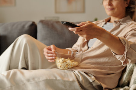 Relaxed woman sitting on sofa with remote control in one hand and a bowl of popcorn in other hand,...