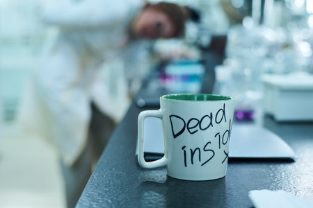 Closeup of mug with dead inside text on it on table in laboratory full of zombie workersの写真素材