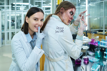 Medium shot of cheerful human female Caucasian lab technician pressing finger to lips after placing sticker on back of zombie colleague, who is studying test tubesの写真素材
