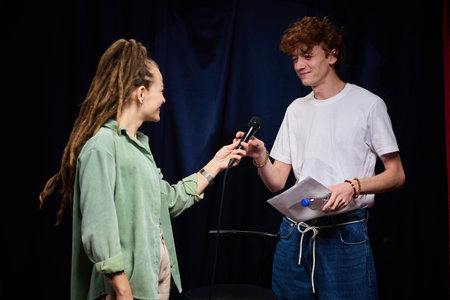 Smiling young man and woman sharing microphone on stage while looking at each other holding paper...