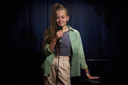 Portrait of smiling woman with long hair, holding microphone, standing on stage. Appearing confident...