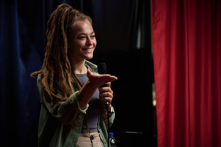 Smiling young woman using microphone and speaking on stage with an audience, standing in front of...
