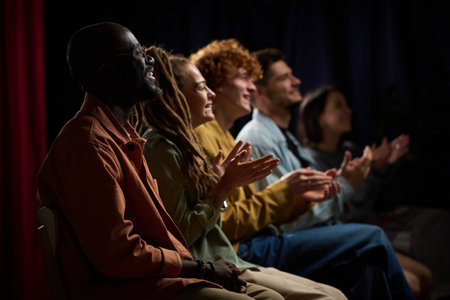 Group of diverse individuals applauding a performance with joyful expressions while seated in a...