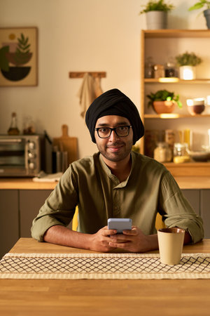 Portrait of smiling man wearing turban and glasses, sitting at wooden table in cozy kitchen, using his smartphone and holding a cup, surrounded by various kitchenware and potted plantsの写真素材