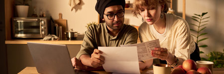 Couple with paper documents sitting at kitchen table with laptop, discussing finances and reading...