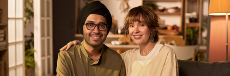 Portrait of smiling multiracial couple sitting side by side in cozy living room, sharing warm moment...