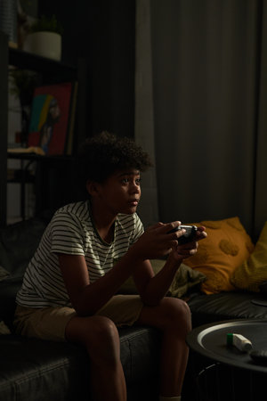 Serious boy in casual apparel pressing buttons on joystick while sitting in front of tv set in domestic room and playing video gameの写真素材