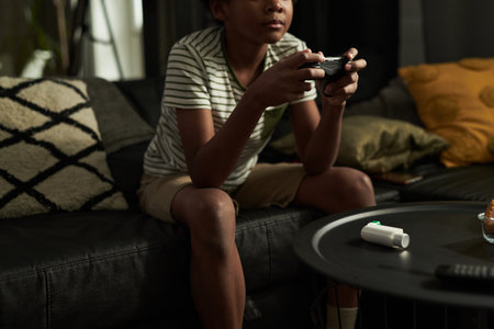 Table with inhaler standing in front of African American child with gamepad sitting on sofa and...