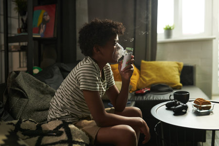 Side view of youthful African American asthmatic boy sitting on couch and having medical treatment