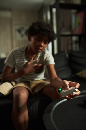 Hand of youthful boy with asthma attack touching his chest and taking inhaler while sitting on couch...