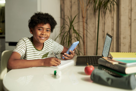 Cute smiling boy in t-shirt looking at camera while using smartphone and taking notes in copybook during distant learningの写真素材