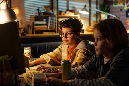 Two children, one wearing glasses, engaging in computer gaming in dimly lit room filled with various...
