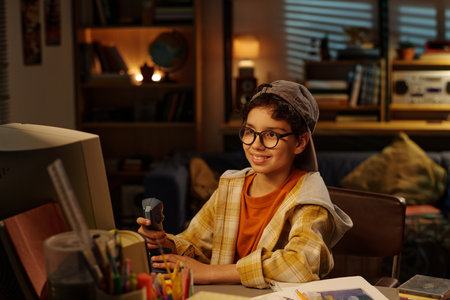 Smiling young boy wearing glasses and a cap, holding walkie talkie while sitting at desk in a cozy,...
