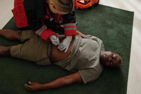 Paramedic providing medical care to injured man lying on green carpet indoors. Medical kit visible...
