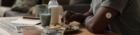Person eating breakfast while monitoring blood glucose levels with a wearable device on arm, with bowls of fresh berries and milk on tableの写真素材