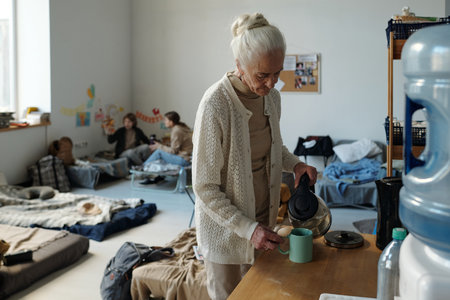 Senior woman in casual attire pouring hot water into mug while standing by table with kitchenware in refugee campの写真素材