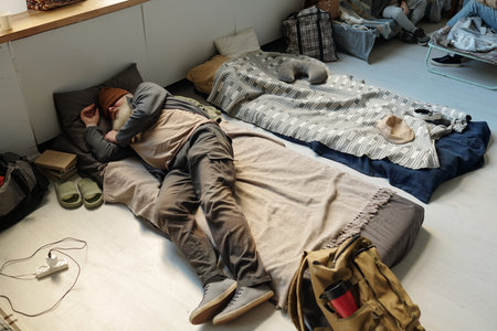High angle of bearded senior man in casual attire sleeping on couchette on the floor of spacious room serving as refugee campの写真素材