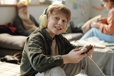 Smiling child in headphones looking at camera while sitting on sleeper and watching online video in...