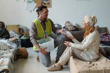 Young volunteer in casualwear and reflective jacket passing bottle of water to senior female refugee...