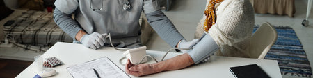 Header of doctor in medical scrubs sitting by table in front of senior female refugee and measuring her blood pressureの写真素材