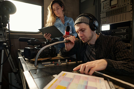 Young male operator adjusting sound on console and speaking in microphone while helping female...