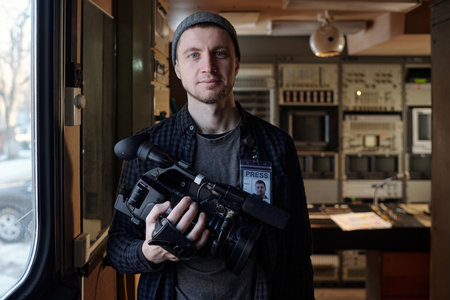 Young successful male operator with professional video camera in hand looking at you while standing...