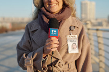 Cropped shot of young smiling female journalist or reporter in beige coat speaking in microphone in...