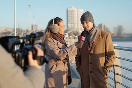 Young man and woman in beige coats standing in front of video camera shooting outdoor interview by riversideの写真素材