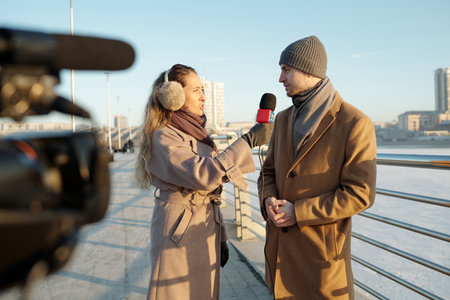 Young female journalist speaking to man in beige coat while holding microphone by his face during outdoor interviewの写真素材