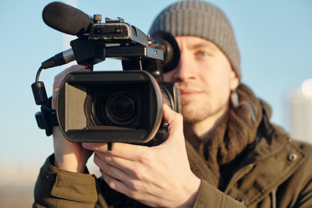 Focus on hand of young male videographer adjusting video camera while preparing for shooting new...