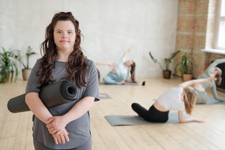 Girl with rolled mat standing on background of females doing yoga exercise on the floorの写真素材