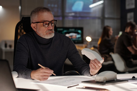 Senior engineer in eyeglasses and pullover sitting by workplace in bureau and looking at car model...