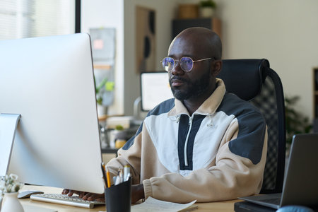 Young serious male designer in eyeglasses and casual attire looking at computer screen and pressing buttons of keypadの写真素材
