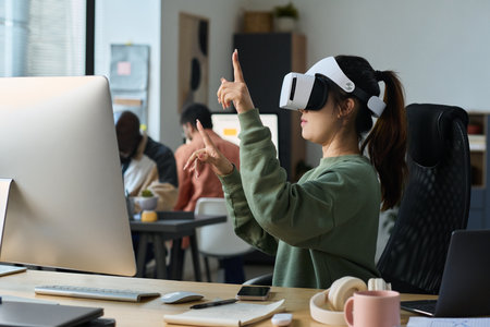 Young Asian businesswoman in VR helmet and casual attire pointing at virtual information while sitting by desk in coworking spaceの写真素材