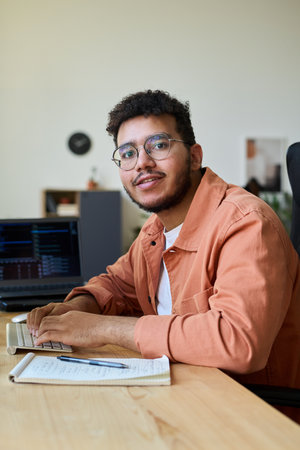 Happy young multiethnic man in eyeglasses looking at camera while typing on computer keyboard by his workplaceの写真素材