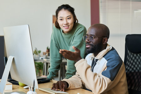 Happy young Asian female programmer standing next to male colleague explaining her points of new project by workplaceの写真素材