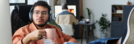 Header of young restful male office worker in headphones having coffee at break and looking at...