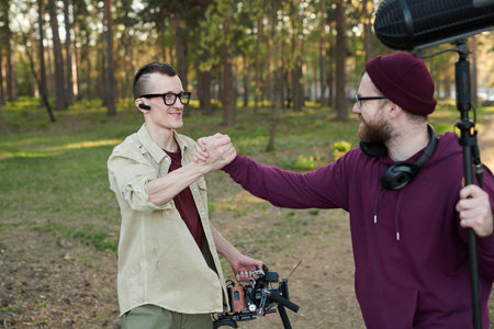 Young cameraman and his assistant with microphone shaking hands in natural environment
