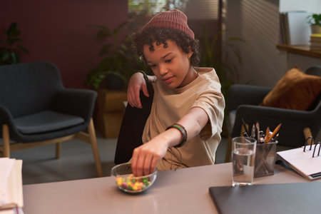 African American schoolchild taking candy from glass bowl while sitting by workplace of school...