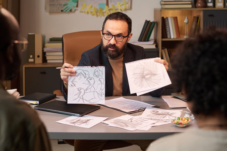 Bearded school psychologist showing child drawings with aggressive symbols to father of schoolboy at therapy sessionの写真素材