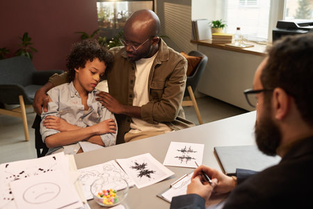School psychologist sitting by desk in front of patient keeping his head on shoulder of his father comforting him at sessionの写真素材