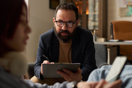 Confident mature male psychologist in eyeglasses looking at patient and taking notes in paper...