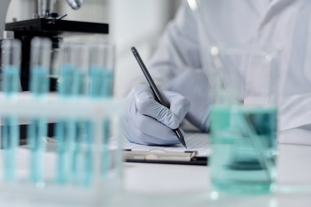 Gloved hand holding pen, interacting with papers, surrounded by laboratory equipment, blue liquid-filled beakers in the foreground and background, indicating focused research environmentの写真素材