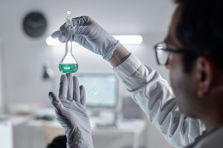 Scientist holding flask with green liquid while meticulously observing analysis in laboratory. Beaker in focus with scientific equipment and computers in backgroundの写真素材