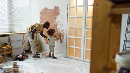 African American father painting wall with young daughter in light-filled room with scattered painting supplies visible around and ladder leaning against wallの写真素材