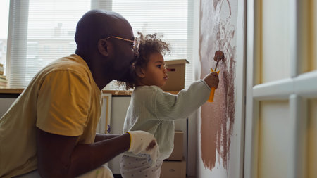 Black person and young child engaging in shared painting activity in cozy room with natural light from large window, creating precious memory togetherの写真素材