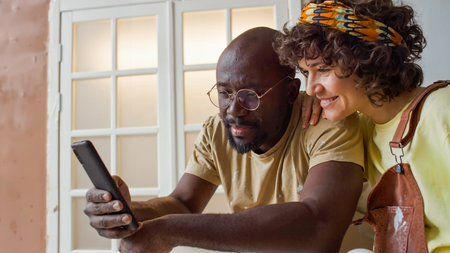 Couple smiling and looking at smartphone inside home with natural lighting coming through window. Man in casual t-shirt and woman in patterned headband both seem engaged with screenの写真素材