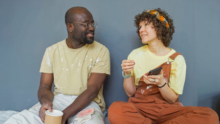 Man and woman sitting on cushions and chatting warmly while holding coffee cups and phone, both...