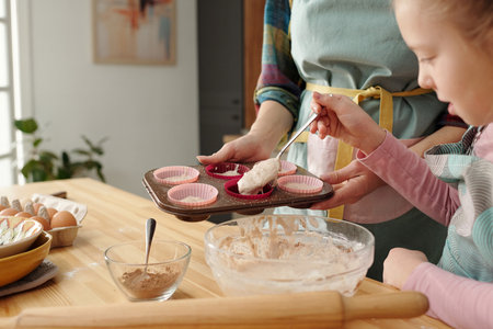 Parent and child baking cupcakes in home kitchen, holding muffin tray with batters next to mixing...