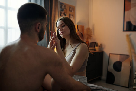 Intimate moment between couple sitting on bed in cozy bedroom, woman gently touching mans face. Warm lighting and decorations in background creating relaxing atmosphereの写真素材